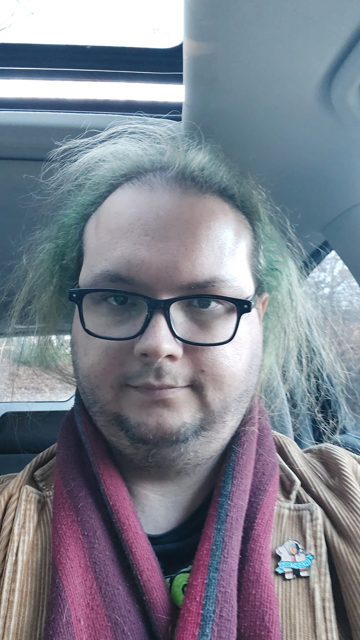 A man with green hair wearing a brown corderoy jacket sits in a car, the sky is cloudy